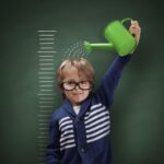 Un enfant avec des lunettes arrose sa tête avec un arrosoir vert devant un tableau noir avec une échelle de croissance dessinée.