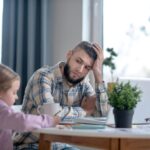 Un père fatigué est assis à une table avec son ordinateur portable, regardant sa fille qui fait ses devoirs.