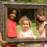 Trois jeunes filles souriantes tiennent un cadre photo en bois dans un parc, créant une composition artistique.