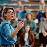 Une conférencière en blouse bleue s’adresse à une audience attentive dans un amphithéâtre.