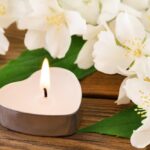 Une bougie en forme de cœur allumée repose sur une table en bois, entourée de fleurs blanches.