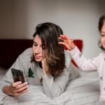 Femme allongée sur un lit, absorbée par son téléphone, tandis qu'un enfant tente d'attirer son attention.