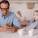 Un homme absorbé par son téléphone portable alors qu’une jeune fille lui tend un cahier.