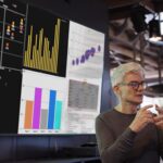 Personne en tenue professionnelle présentant des statistiques et des graphiques sur un grand écran.