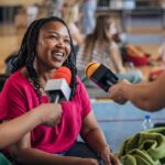 Femme souriante interviewée par plusieurs journalistes avec des micros colorés.
