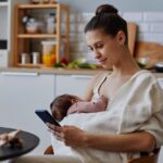 Une jeune mère allaite son bébé tout en consultant son smartphone dans une cuisine moderne et chaleureuse.