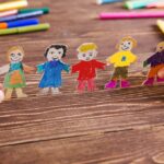 Deux mains tiennent une guirlande de personnages en papier colorés, représentant des enfants souriants, sur une table en bois entourée de crayons.