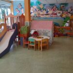 Salle de jeux colorée d'une crèche avec toboggan en bois, coin lecture, fresque murale animée et mobilier adapté aux enfants.